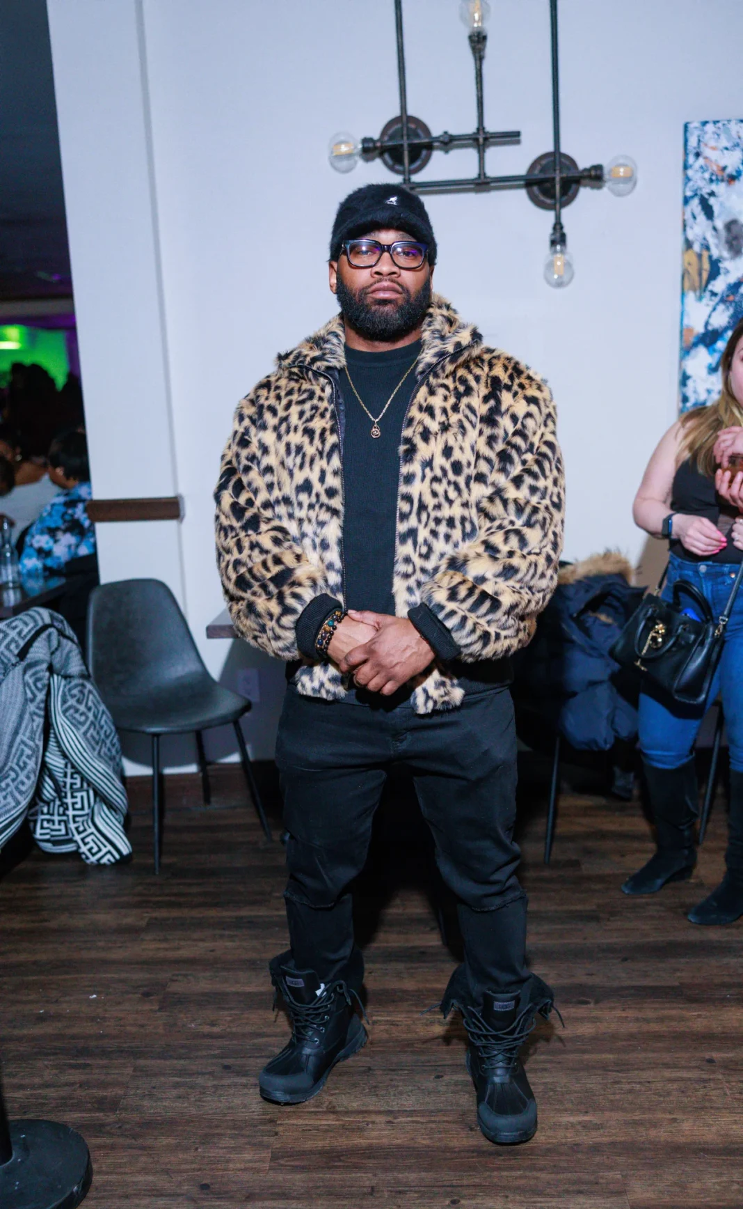 SALT + KO 2-22-25 Saturday A fashionable man in a leopard-print coat posing at Salt & Ko in Southfield, Michigan, with a vibrant nightlife atmosphere in the background.
